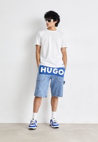 Camisa de algodón blanca combinada con pantalones cortos de mezclilla azules con un gran gráfico de "HUGO", complementada con zapatillas azules y blancas.