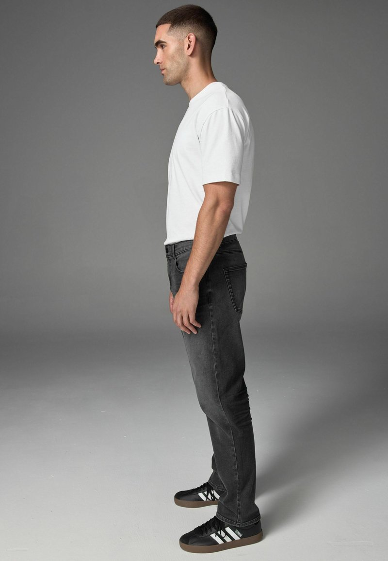 Weißes Baumwoll-T-Shirt, dunkelgraue Slim-Fit-Jeans und schwarze Sneakers mit weißen Streifen. Das Modell wird im Profil vor einem schlichten Hintergrund gezeigt.