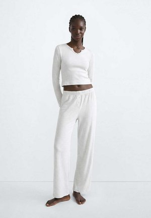 Woman standing barefoot wearing a white long-sleeve crop top and loose white pants against a plain light background.