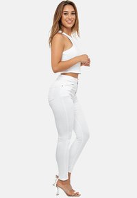 Jeans ajustados blancos con cintura alta, silueta slim y detalles notables en los bolsillos. Combinados con una camiseta sin mangas blanca corta y tacones transparentes.