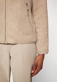 Giacca in pile beige con superficie texturizzata e rifiniture contrastanti color nocciola. Tasca con zip a sinistra; logo ricamato in basso.