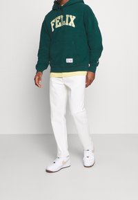 Felpa verde con "FELIX" in lettere gialle, tasca a canguro, indossata sopra una maglietta gialla, abbinata a pantaloni bianchi e sneakers bianche.