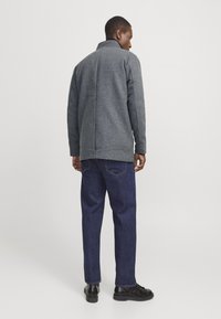 Homme portant une veste grise, un jean bleu foncé et des chaussures noires, se tenant de dos contre un fond clair.