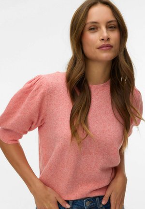 Kobieta o długich brązowych włosach, ubrana w różowy sweter z bufkami i niebieskie dżinsy, stoi z rękami w kieszeniach na białym tle.