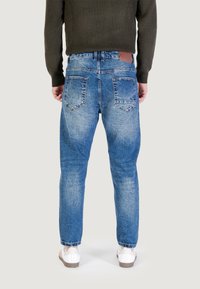 Uomo con jeans blu sbiaditi e sneaker bianche, in piedi con la schiena rivolta contro uno sfondo chiaro e semplice.