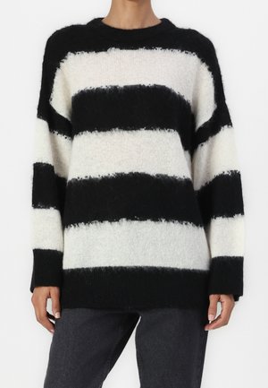 Osoba ubrana w czarno-biały, miękki sweter w szerokie poziome pasy oraz ciemne spodnie na jednolitym tle.