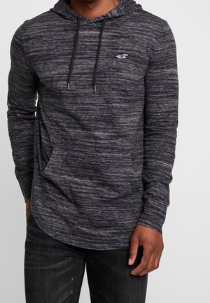 Mann trägt dunkelgraues Kapuzensweatshirt mit langen Ärmeln, dezentem horizontalem Marmorierungsmuster und Kängurutasche vorne, schwarze Hose.