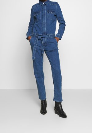 Persoon draagt een blauwe denim jumpsuit met lange mouwen, voorzakken, een tailleband en zwarte enkellaarzen, staat met handen in de zakken.
