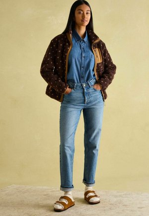 Brown fleece jacket with abstract pattern, denim button-up shirt, light blue jeans, brown sandals with white straps, textured background.