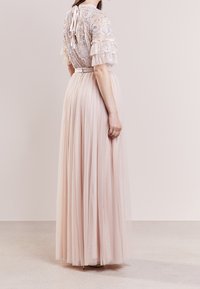 Lange, pastelroze jurk met een doorzichtig borduursel bovenstuk, volants mouwen, een ceintuur op de taille en een vloeiende tule rok. Achteraanzicht getoond.