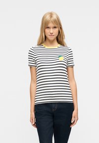 Νεαρή γυναίκα που φοράει ριγέ λευκό και μαύρο κοντομάνικο πουκάμισο με κίτρινο μοτίβο λεμονιού και κίτρινο γιακά, συνδυασμένο με σκούρο μπλε τζιν παντελόνι.