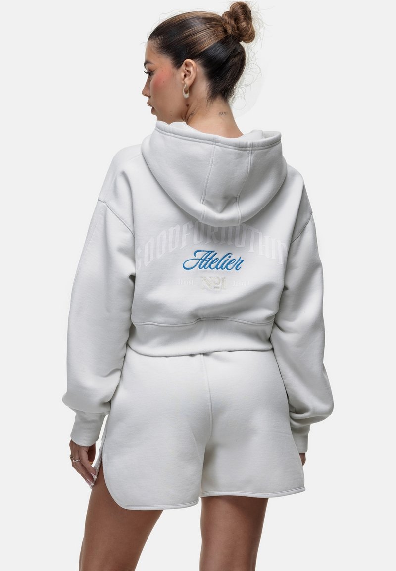 Felpa cropped bianca con cappuccio, caratterizzata da scritte sul retro e pantaloni corti bianchi abbinati. Tessuto morbido e testurizzato con vestibilità rilassata.