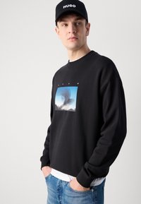 Felpa nera con maniche lunghe, presenta una stampa grafica centrale in blu. Abbinata a un cappellino HUGO e indossata con jeans azzurri chiari.