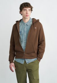 Καφέ φούτερ με φερμουάρ, κατασκευασμένο από απαλό ύφασμα, με μπροστινή τσέπη, κουκούλα με κορδόνι και λογότυπο, φορεμένο πάνω από τζιν πουκάμισο και πράσινο παντελόνι.