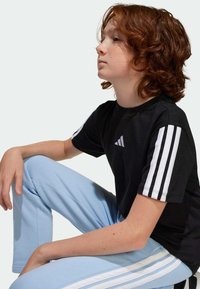 T-shirt en coton noir avec des détails en trois rayures blanches sur les manches et un logo gris. Pantalon bleu clair avec des motifs de rayures blanches sur les côtés.