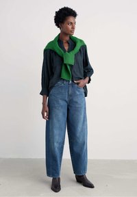 Žena oblečená v tmavej kockovanej blúzke, modrých širokých džínoch, čiernych čižmách a so zeleným sveterom prehodeným cez plecia, stojí na svetlej podlahe.