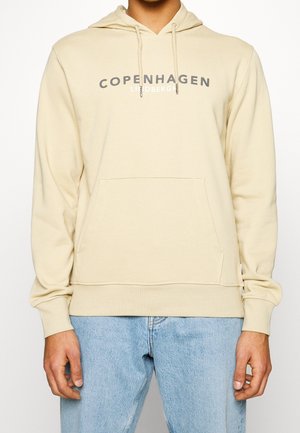 Person trägt beige Kapuzenpullover mit dem Schriftzug "COPENHAGEN" und hellblaue Jeans, zeigt Oberkörper und Arme vor einfachem weißem Hintergrund.