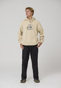 Beige hoodie with a front pocket and graphic print. Paired with black pants and camouflage shoes. Plain background.