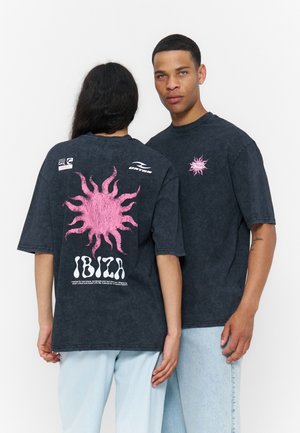 Dvaja ľudia majú na sebe tmavé oversized tričká s ružovými slnečnými motívmi a textom „IBIZA“, jeden smeruje smerom dopredu, druhý ukazuje grafiku na chrbte.