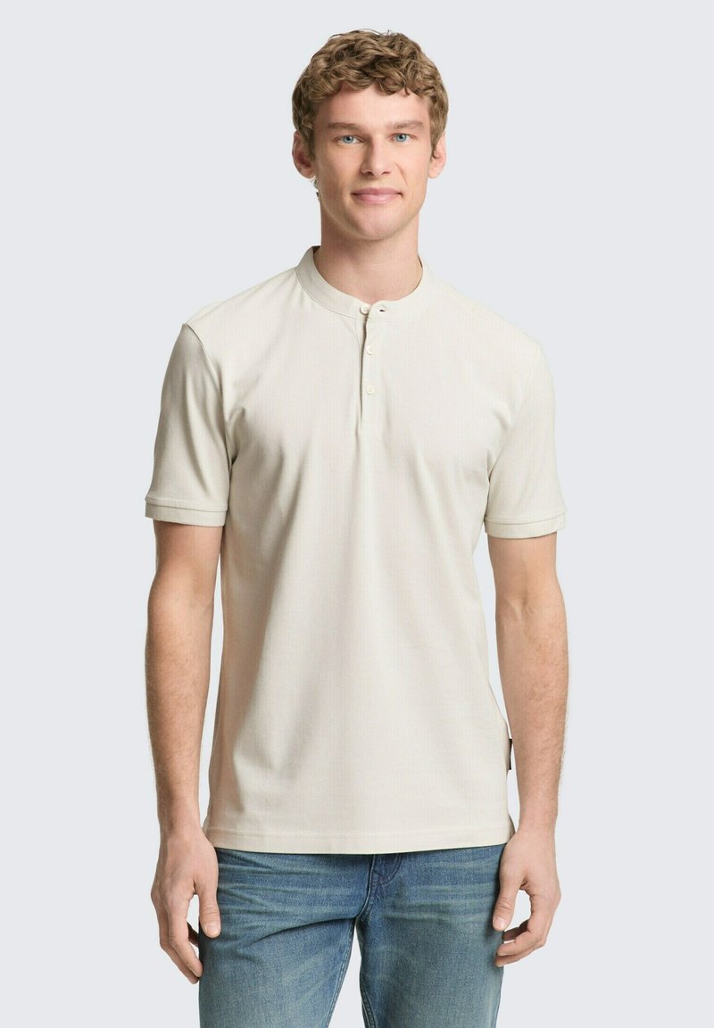 Giovane con capelli ricci che indossa una maglietta henley cremosa a maniche corte e jeans blu, in piedi contro uno sfondo chiaro e uniforme.