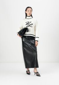 Pull blanc à rayures avec inscriptions noires, jupe crayon en cuir noir, et mules à talons noirs avec des accents à clous. Modèle tenant une pochette.