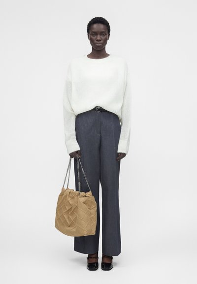 Γυναίκα που φοράει άσπρο πουλόβερ και γκρι ψηλόμεσο παντελόνι, κρατώντας μία μπεζ υφασμάτινη τσάντα με αλυσίδα, στέκεται σε λευκό φόντο.