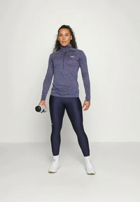 Donna in piedi, che indossa una maglietta sportiva lunga viola e leggings scuri, mentre tiene in mano destra una bottiglia d'acqua bianca.