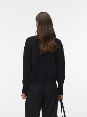 Kvinde med langt brunt hår iført en sort strikket sweater og sorte bukser, stående med ryggen til, holder en sort rem i højre hånd.