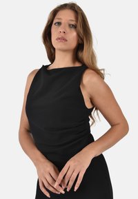 Abito nero senza maniche con una silhouette aderente, texture liscia e un delicato scollo a barca; la modella posa con le mani sui fianchi.