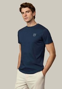 Joven con cabello castaño vistiendo una camiseta Hackett azul marino y pantalones crema, de pie con las manos en los bolsillos frente a un fondo beige.