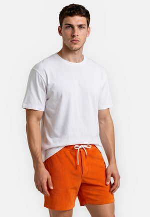 Ung mand iført en plain hvid T-shirt og lyse orange shorts med snøre mod en hvid baggrund.