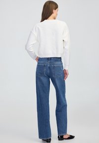 Kobieta z długimi brązowymi włosami, ubrana w biały sweter w kropki, niebieskie proste dżinsy i czarne buty, stojąca tyłem do aparatu.