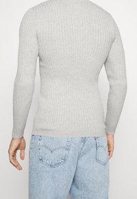 Maglietta a maniche lunghe grigio chiaro a coste con un design aderente, scollo rotondo e texture liscia. Indossata con jeans denim azzurri caratterizzati da tasche posteriori.