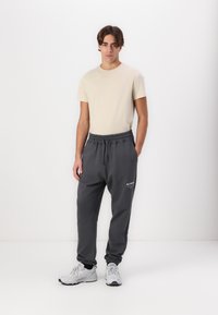 Beige T-Shirt und graue Jogginghosen. Die Hosen verfügen über einen elastischen Taillenbund und ein Logo-Detail. Das Modell trägt graue Sneaker mit einem strukturierten Design.
