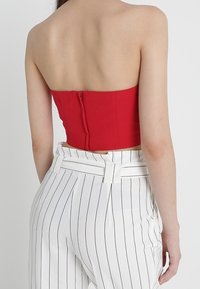 Kvinna iklädd en axelbandslös röd topp med dragkedja bak och högtalande vita byxor med svarta vertikala randningar, sedd från baksidan.