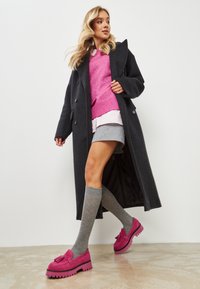 Langer schwarzer Mantel, rosa gestrickter Pullover, grauer Rock, graue kniehohe Socken und rosa Slipper mit dicken Sohlen, kombiniert mit schichtartigen Texturen.