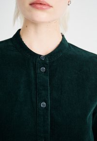 Chemise en velours côtelé vert foncé avec un col montant, présentant des boutons noirs et une texture côtelée. Gros plan sur les détails du haut du vêtement et du col.