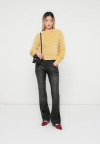 Jongeman die staat in een gele fuzzy trui, zwarte high-waist broek, rode ballerina's en een kleine zwarte crossbody tas tegen een witte achtergrond.