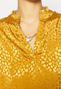 Gul blus i ett glansigt, strukturerat tyg med ett prickigt mönster och en volangkrage. Inkluderar en kedja med detaljer i form av ett hänglås.