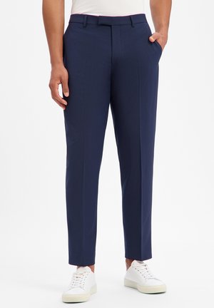 Mann trägt schlanke marineblaue Hose mit vorderen Bügelfalten und weiße Turnschuhe, steht mit einer Hand in der Tasche.