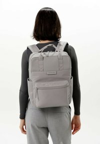 Zaino grigio con spallacci imbottiti, tasca frontale con zip e tasche laterali, realizzato in materiale resistente, con un design strutturato.