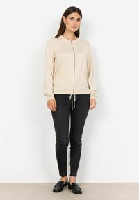 Cardigan beige con zip e maniche lunghe, due tasche frontali, orlo con coulisse e polsini a costine, abbinato a jeans skinny neri e mocassini.