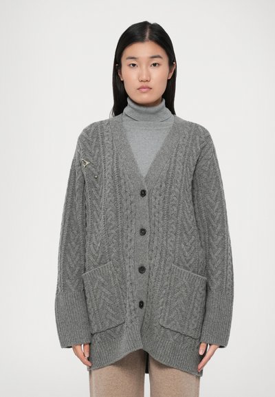 Jeune femme portant un cardigan en tricot torsadé gris sur un pull col roulé gris et un pantalon beige, se tenant devant un fond uni.
