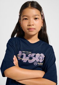 Mörkblå t-shirt i bomull med trycktexten "SERVE THE GAME" i ljuslila och guld, med avslappnad passform och korta ärmar.