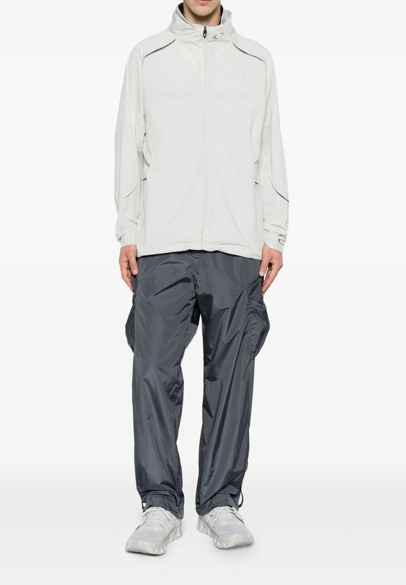 Uomo che indossa una giacca leggera grigio chiaro con zip, pantaloni cargo grigio scuro e scarpe da ginnastica bianche, in piedi con le mani lungo i fianchi.
