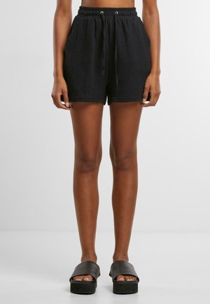 Person iført sorte shorts med tekstureret elastisk talje med snøre og sorte platformsandaler, stående mod en ensfarvet baggrund.