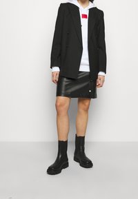 Blazer negro de doble botonadura sobre una sudadera blanca con un acento rojo, combinado con una falda de cuero negra y botines negros. Texturas suaves y diseño elegante.