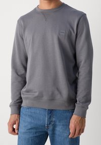 Szary sweatshirt wykonany z miękkiego materiału, z okrągłym dekoltem i ściągaczami przy rękawach. Mała naszywka z logo znajduje się na lewej stronie klatki piersiowej.