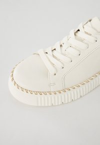 Witte leren sneaker met ronde neus, textuur op het bovenwerk, platte vetersluiting en een gouden touwaccent langs de zool. Stevige witte zool.