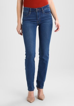 Vrouw draagt blauwe slim-fit jeans, rode mouwloze top en beige hoge hakken, staand op een effen lichte achtergrond.